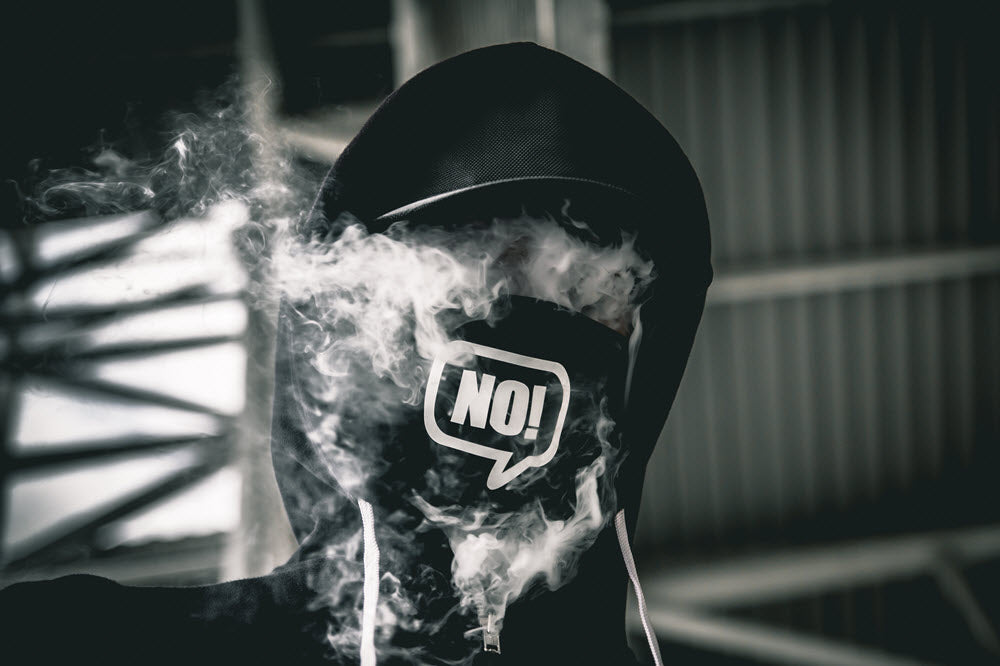 Photo by Rist Art on Unsplash - Black and White photo of hooded person with smoke disguising face and a bandanna covering mouth that reads 'No'